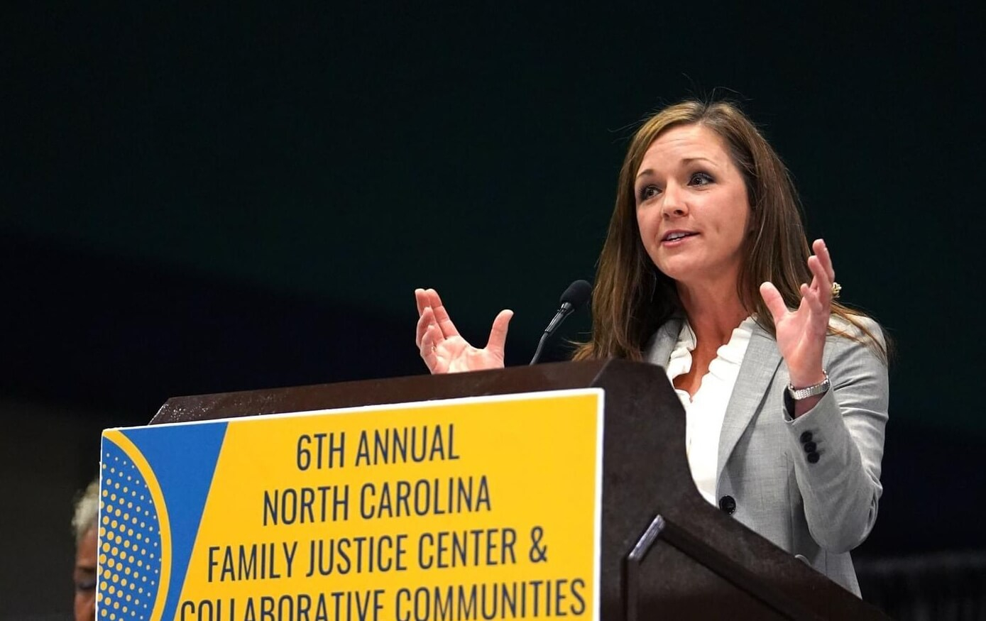 A woman speaking at a podium with a black background, using her hands to communicate body language.