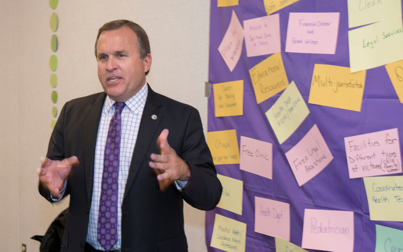 Casey Gwinn of Alliance for HOPE speaking in front of a wall covered with colorful sticky notes featuring topics like victim support facilities.